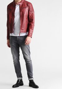 Rode leren jas met een hoge kraag, rits aan de voorkant en gegroefde schouderaccenten. Gecombineerd met grijze jeans en zwarte enkellaarsjes.