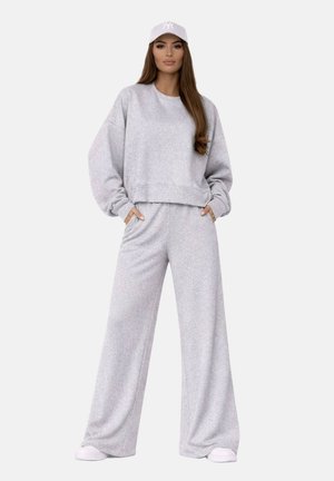 Femme debout les mains dans les poches, portant un sweat-shirt ample gris clair assorti et un pantalon large, avec une casquette blanche et des baskets.