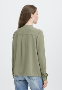 Femme aux longs cheveux bruns portant un blouse à manches longues vert olive et un jeans en denim bleu clair, tournée dos à l'appareil photo.