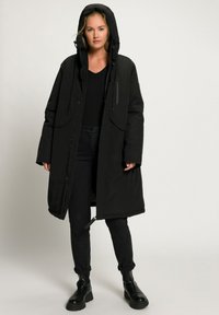 Parka noir long avec capuche, coutures d'épaules, deux poches avant et fermeture éclair. Porté sur une chemise noire et un pantalon noir.