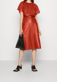 Haut en cuir rouge avec encolure froncée, jupe midi en cuir rouge assortie avec ceinture, sac triangular noir et sandales à talons noires.