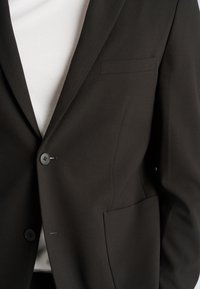 Zbliżenie na czarną, dopasowaną marynarkę z dwoma guzikami, kieszenią piersiową z wypustką oraz boczną kieszenią z patką, noszoną na białej koszuli.