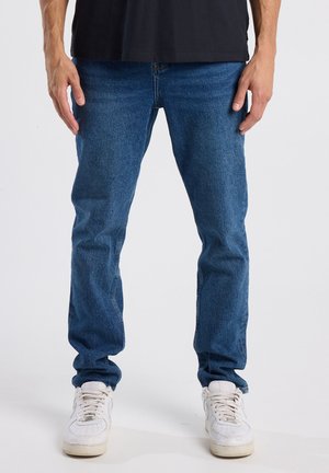 Jean en denim bleu à coupe droite, avec une taille mi-haute et un design classique à cinq poches. Porté avec des baskets blanches.
