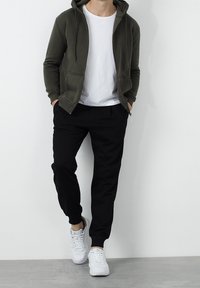Sudadera con cremallera color verde oliva, camiseta blanca, pantalones negros ajustados y zapatillas blancas. Tejido suave y corte informal con puños acanalados.