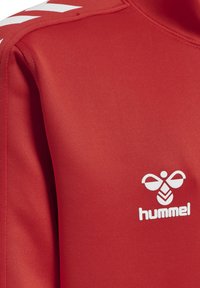 Giacca sportiva rossa realizzata in tessuto liscio, caratterizzata da un logo bianco con un design di ape e da contrasti di chevron bianchi sulle spalle.