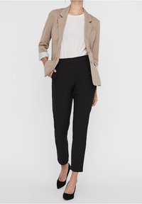 Žena má na sobě béžový blazer přes bílý top, černé kalhoty na míru a černé podpatky, stojí s jednou rukou v kapse na bílém pozadí.