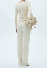 Pull ajusté beige clair à manches longues et col large, assorti à un pantalon à jambes larges de la même couleur. Le modèle tient un sac beige.