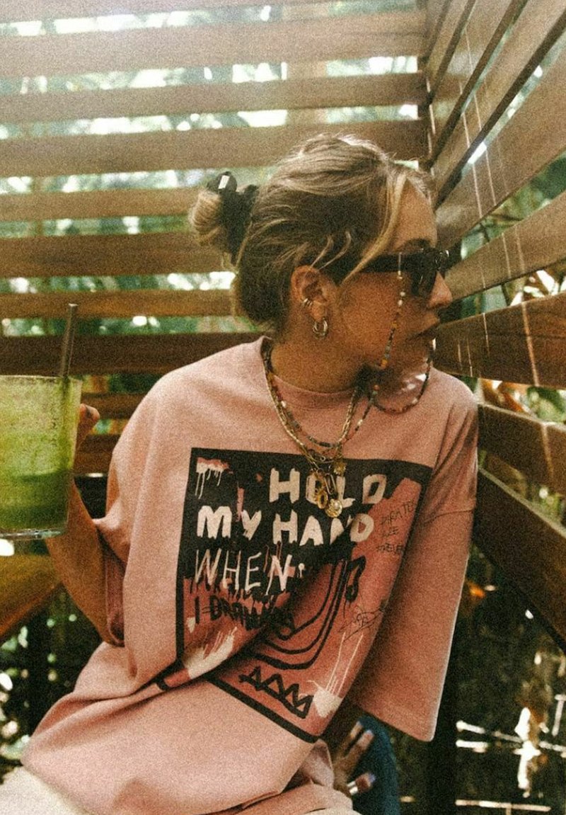 Różowa, oversize'owa koszulka z grafiką, z tekstem i spływającymi detalami. Dodatki to kilka złotych naszyjników i okulary przeciwsłoneczne.