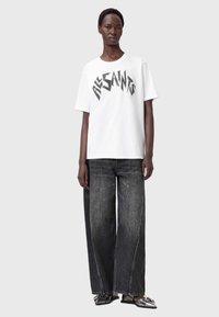 T-shirt bianca a maniche corte con grafica nera "ALLSAINTS", abbinata a jeans a gamba larga in denim scuro e scarpe con accenti metallici.
