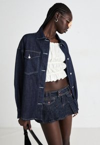 Denim jacket with a front pocket, white ruffled crop top, and denim miniskirt. Dark blue fabric with contrasting stitching and button accents.