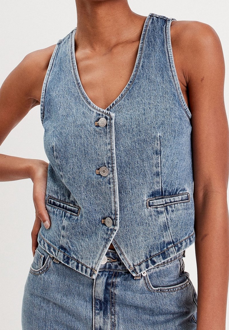 Personne portant un gilet en denim bleu clair et un jean assorti, la main reposant sur la hanche devant un fond uni.