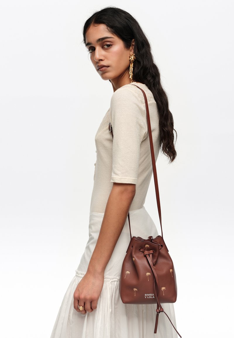 Pikajuustega naine kannab kreemika värvi topi ja seelikut, kuldseid ripatavaid kõrvarõngaid ning tal on käes pruunatsükliline kott palmileheprintidega.