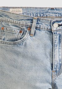 Světle modré džíny Levi's zblízka, ukazující přední kapsu, poutko na opasek a kovový knoflík s detaily šití.