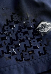 Tissu bleu foncé avec un motif de texte répétitif brillant "J90" et un patch cousu indiquant "Jack & Jones Authentic Denimwear Since 1990."