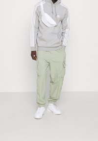 Hombre vestido con sudadera gris de Adidas con rayas blancas, pantalones cargo verde claro, zapatillas blancas y una bolsa cruzada blanca, contra un fondo liso.