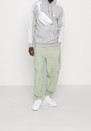 Muž v šedé mikině Adidas s bílými pruhy, světle zelených cargo kalhotách, bílých teniskách a bílé crossbody tašce na neutrálním pozadí.