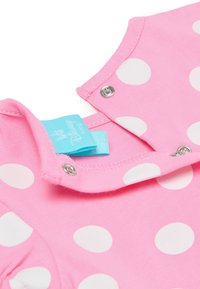 Pink cotton onesie featuring large white polka dots, with a snap closure at the collar and a blue brand tag. Smooth texture.