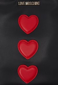 Černý kožený povrch s třemi vertikálními červenými prošitými srdci a zlatým textem "LOVE MOSCHINO" nahoře.