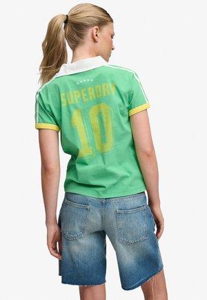 Lyshåret kvinde med hestehale iført grøn Superdry polo-shirt med gult nummer 10 og falmede denimshorts, vendt væk fra kameraet.