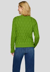 Grüner Strickpullover mit strukturiertem Muster und geripptem Saum. Getragen über blauer Jeans mit sichtbaren Gesäßtaschen.