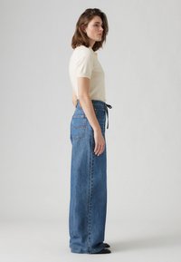 Kreem värvi lühikeste varrukatega kudum, millele on paired kõrge vööjoonega sinised denimpüksid, laiad sääred. Pükstel on vöö peal side ja tagapooltaskute õmblused.