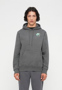 Jovem com cabelo encaracolado escuro, vestindo um hoodie cinza da Nike Air e calças cinzas a condizer, em pé contra um fundo branco simples.