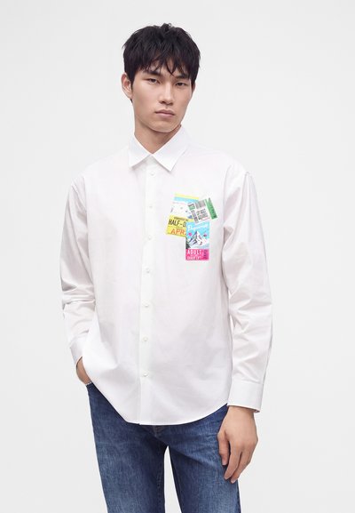 Jeune homme portant une chemise blanche à boutons avec des motifs colorés de billets sur la zone de la poche poitrine et un jean bleu, debout devant un fond blanc.