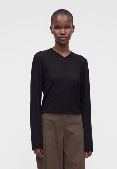 Femme aux cheveux courts portant un haut noir à manches longues et col en V ainsi qu'un pantalon marron, debout devant un fond clair uni.
