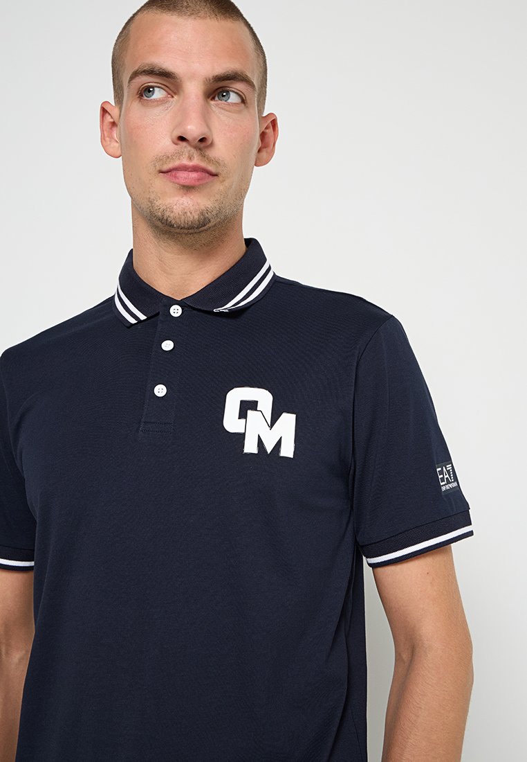 Granatowa koszulka polo z białymi akcentami, z trzema guzikami z przodu, oraz dużym haftowanym logo "CM" na piersi. Krótkie rękawy z paskowanymi mankietami.