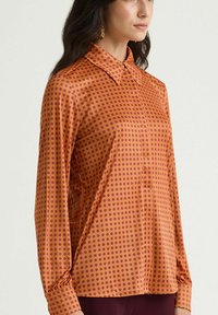 Camicia a maniche lunghe in raso arancione con pois viola, con collo a risvolto e chiusura con bottoni, polsini arricciati e vestibilità comoda.