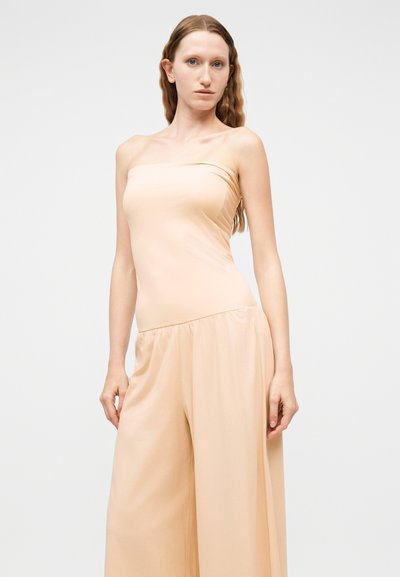 Γυναίκα με μακριά κυματιστά μαλλιά, φορώντας ένα μπεζ ολόσωμο κορμάκι χωρίς τιράντες, όρθια με τα χέρια χαλαρά στα πλευρά της, μπροστά σε απλό φόντο.