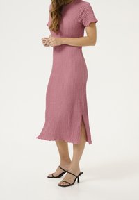Strick-Kleid mit kurzen Ärmeln in Midi-Länge, in Pink, mit strukturiertem Muster und seitlichem Schlitz; kombiniert mit schwarzen Riemchenschuhen.