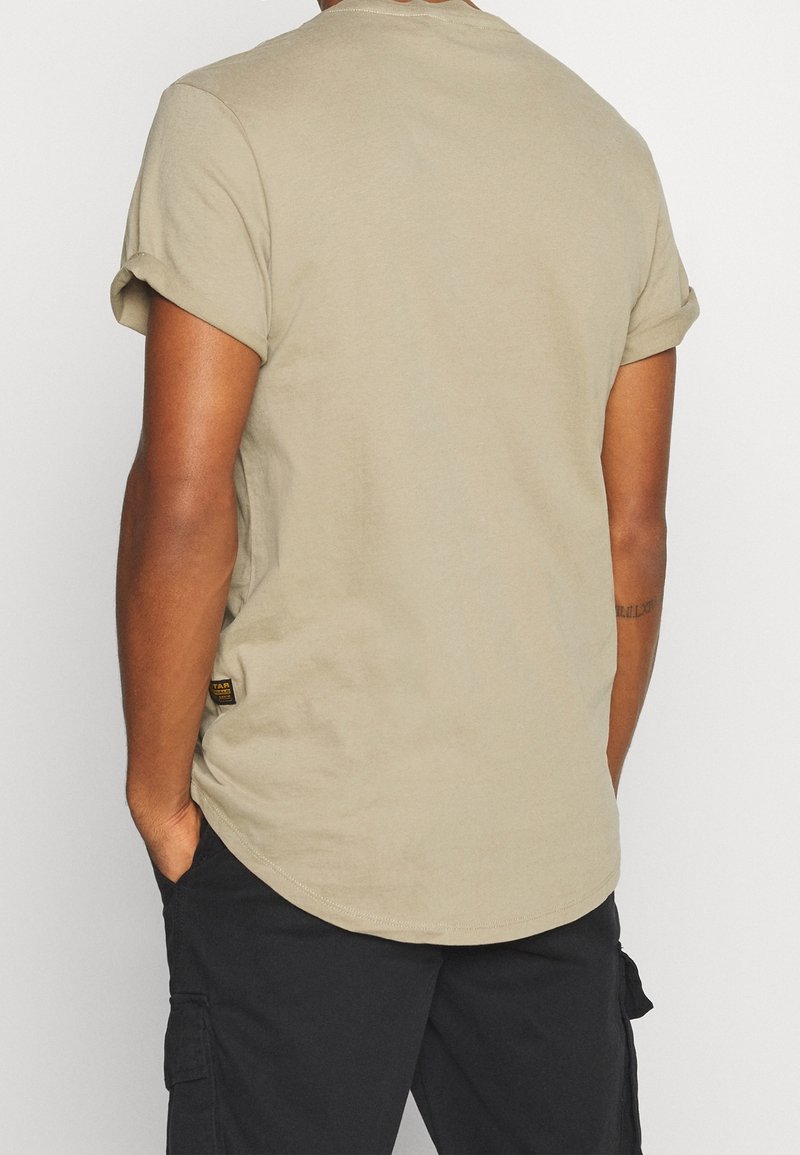 Homme portant un T-shirt beige à manches courtes avec revers retroussés et un short cargo noir, les mains dans les poches, dos à l'appareil photo.
