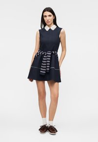 Femme portant une robe sans manches bleu marine avec un col en dentelle blanche, un pull rayé noué à la taille, des chaussettes blanches et des chaussures marron à pompons.