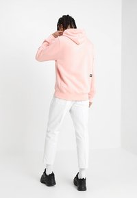 Man med flätat hår som står, iförd en rosa hoodie, vita byxor, vita strumpor och svarta sneakers mot en vit bakgrund.