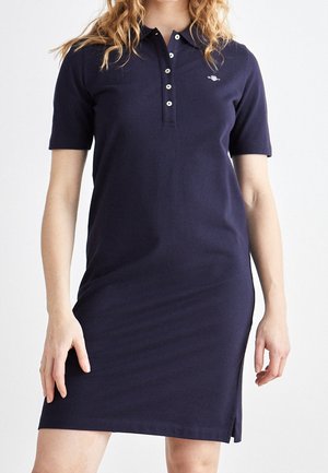 Nainen pukeutuneena tummansiniseen lyhythihaiseen polomekkoon, jossa on nappilista ja pieni logo vasemmassa rinnassa, seisoo yksinkertaisen taustan edessä.