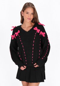 Schwarzer Pullover mit V-Ausschnitt, rosa Schleifendetails an den Schultern und kontrastierendem rosa Stich entlang der Vorderseite. Weicher, strukturierter Stoff.