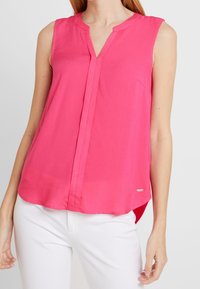 Blouse sans manches rose avec un col en V, tissu doux, patte de boutonnage à l'avant et ourlet arrière légèrement plus long. Marque discrète en bas.