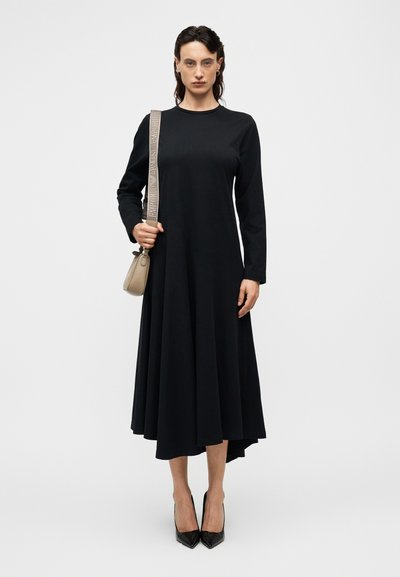Μαύρο φόρεμα με μακριά μανίκια και φαρδιά γραμμή στο τελείωμα, φτιαγμένο από απαλό ύφασμα. Συνδυάζεται με μικρή μπεζ τσάντα και μαύρες μυτερές γόβες.