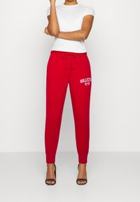 Röda sweatpants med dragsko i midjan, med "HOLLISTER"-logotyp i vitt. Bärs med en vanlig vit t-shirt och svarta högklackade sandaler.