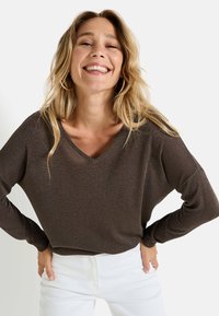 Suéter marrón de cuello en V con un acabado suave y texturizado. Mangas largas y ajuste relajado. Pantalones blancos visibles en la cintura, creando un contraste.