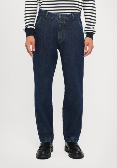 Dunkelblaue Jeans aus Denim mit lässigem Schnitt, hoher Taille, Ein-Knopf-Verschluss und sichtbaren Nähten. Getragen mit einem gestreiften langärmligen Oberteil.