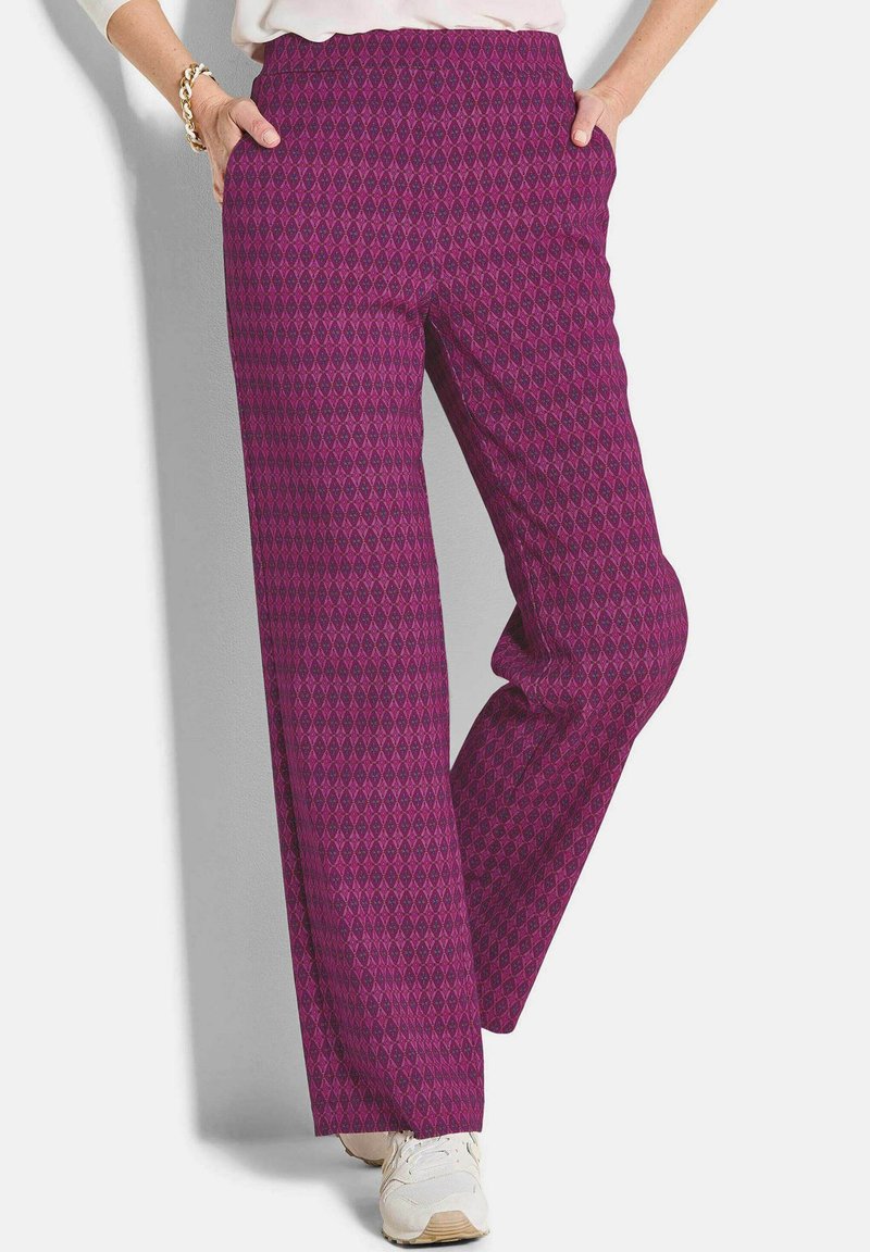 Vrouw in paarse, gebloemde wijde broek en witte sneakers, met handen in haar zakken, tegen een effen lichte achtergrond.