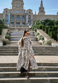 Leopárdmintás műszőrme kabát széles gallérral, övvel a derékon, és hosszú ujjakkal, áttetsző fekete csipkebebújós ruha fölött, fűzős magassarkú cipővel viselve.