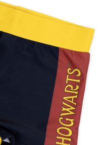 Zwemshorts met een gele tailleband, een marineblauwe body en een kastanjebruin paneel met "HOGWARTS" in gele tekst gedrukt. Glad stoftextuur.
