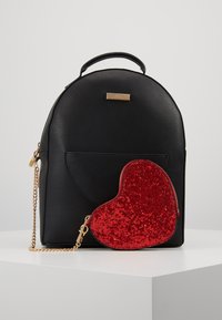 Sac à dos en similicuir noir à forme arrondie, doté d'une poche avant. Un porte-monnaie rouge en forme de cœur pailleté avec une fermeture éclair dorée y est attaché.