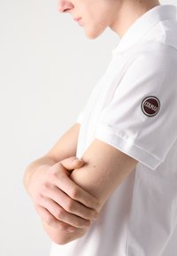Balta polo krekls ar īsām piedurknēm, kas rotāts ar apaļu Colmar logo uz kreisās piedurknes. Tuvplāns, izceļot roku stāvokli.