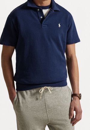 Mann trägt ein dunkelblaues Polohemd mit weißem, besticktem Logo, darüber ein schwarz-weißes Shirt, kombiniert mit hellgrauen Jogginghosen mit Kordelzug.