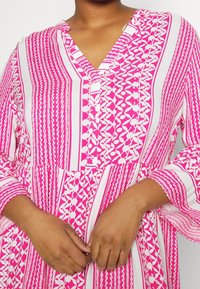 Vestido con estampado en rosa y blanco, con escote en V, mangas largas y una silueta fluida. Presenta diseños geométricos y florales. Tejido ligero.