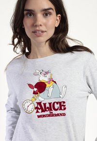 Mujer joven con una camiseta gris clara que presenta al Conejo Blanco sosteniendo un reloj de bolsillo y el texto "Alicia en el País de las Maravillas."